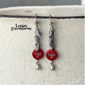 May include: A pair of dangle earrings featuring red, round beads with a bee design, silver-colored leaf accents, and teardrop-shaped dangles. The earrings are suspended from silver-colored hooks. The text "Leslie's Expressories" is visible.