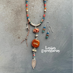 Peut inclure: Un long collier de perles multicolores avec un pendentif en argent orné d'un charme de girafe, d'une perle en céramique brune à imprimé girafe, d'une perle en céramique brune avec le mot "Love" inscrit dessus et d'un charme en feuille d'argent. Le collier est assorti à des boucles d'oreilles assorties avec des perles brunes et bleues.