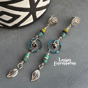 May include: A pair of dangle earrings with silver-toned leaf charms and turquoise beads. The earrings feature a silver-toned flower charm within a blue ring, with yellow and green beads. The earrings are on a gray surface.