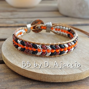 Op de afbeelding: Een handgemaakte armband met zwarte, oranje, zilveren en bordeauxrode kralen. De kralen zijn in rijen gerangschikt en bevestigd aan een bruine leren band. De armband heeft een knoopsluiting en de tekst "BB by D. Ajaccio".
