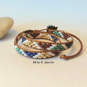 Puede incluir: Un brazalete de cuero envuelto con cuentas multicolores con un dije de sol. El brazalete está hecho con cuentas blancas, amarillas, verdes, azules y rojas. El texto "BB by D. Ajaccio" es visible en la superficie blanca.