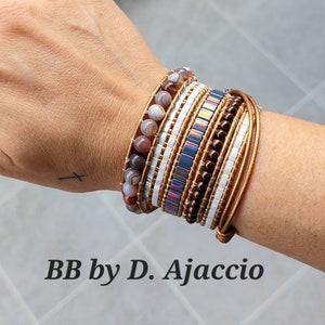 Puede incluir: Un conjunto de pulseras en la muñeca de una persona. Las pulseras están hechas de diferentes materiales, incluyendo cuero, cuentas y piedras. Las pulseras son en tonos de marrón, blanco y morado. El texto "BB by D. Ajaccio" es visible debajo de las pulseras.