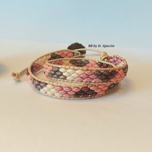 Puede incluir: Un brazalete de cuentas multi-hilo con cuentas de color rosa, blanco y marrón. El brazalete está envuelto alrededor de un cordón de cuero con un cierre de metal. El texto "BB by D. Ajaccio" es visible en el brazalete.