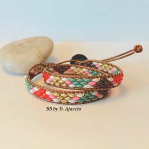 Op de afbeelding: Een veelkleurige kralen armband met een bruine leren wikkel. De armband heeft rode, roze, witte, gele, groene en bruine kralen. De leren wikkel heeft een knoopsluiting. De armband is gelabeld "BB by D. Ajaccio".