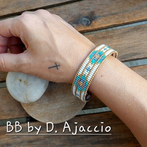 Op de afbeelding: Een witte en turquoise kralen armband met gouden accenten. De armband zit om de pols van een persoon. De tekst "BB by D. Ajaccio" is zichtbaar onder de armband.