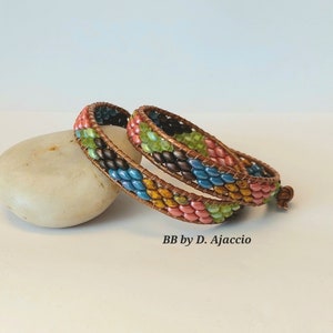 May include: Two beaded bracelets with a chevron pattern in shades of brown, pink, green, blue, and yellow. The bracelets are wrapped around a light-coloured stone. The text "BB by D. Ajaccio" is visible below the bracelets.
