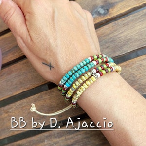May include: A stack of beaded bracelets in turquoise, yellow, red, and green. The bracelets are wrapped around a wrist. The bracelets are made of small beads and silver-colored spacers. The text "BB by D. Ajaccio" is at the bottom.