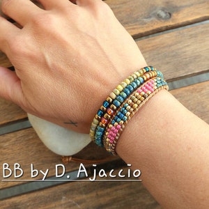 Pode incluir: Um conjunto de quatro pulseiras de contas em tons de azul, verde, dourado e rosa. As pulseiras estão sobrepostas no pulso de uma pessoa. O texto "BB by D. Ajaccio" é visível na parte inferior da imagem.