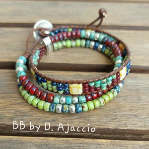 May include: Wrap bracelet with colorful beads in shades of red, green, blue, and yellow. The bracelet has a brown leather band and a silver button clasp. The text "BB by D. Ajaccio" is visible.