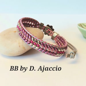 Op de afbeelding: Een roze en gouden kralen armband met een zilveren sluiting. De armband ligt op een lichtbruine steen. De tekst "BB by D. Ajaccio" staat onder de armband.