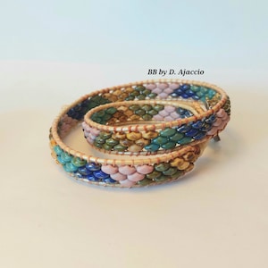 Op de afbeelding: Twee kralenarmbanden met een geweven ontwerp in tinten roze, blauw, groen en bruin. De armbanden zijn om elkaar heen gewikkeld en hebben een koperen draadaccent. De tekst "BB by D. Ajaccio" is zichtbaar in de afbeelding.