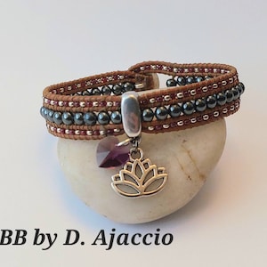Op de afbeelding: Een wikkelarmband met bruin leer, donkergrijze kralen en kleine rode kralen. Een zilveren bedel met een paarse kristal en een lotusbloem hangt aan de armband. De tekst "BB by D. Ajaccio" staat onderaan.