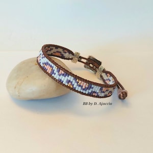 May include: A brown leather bracelet with a white, pink, and purple beaded pattern. The bracelet has a silver clasp and a sliding knot.