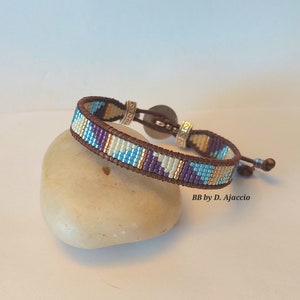 Puede incluir: Un brazalete de cuero marrón con un patrón de cuentas en tonos de morado, azul, blanco y dorado. El brazalete tiene un cierre plateado con el número 2022 grabado en él. El brazalete está descansando sobre una roca marrón claro. BB by D. Ajaccio está impreso en la roca.