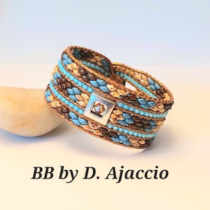 Peut inclure: Un bracelet multi-rangs en perles de couleur marron, beige et bleu. Le bracelet est doté d'un fermoir argenté avec un cristal transparent au centre. Le bracelet est posé sur une surface blanche avec un rocher brun en arrière-plan. BB by D. Ajaccio.