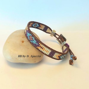 Könnte beinhalten: Ein Armband mit Perlen, braunem Lederband und silberfarbenem Verschluss. Das Armband zeigt ein geometrisches Muster in Lila, Türkis, Gold und Creme. Der Text "BB by D. Ajaccio" ist sichtbar.