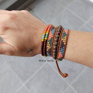 Pode incluir: Um conjunto de seis pulseiras de contas coloridas com fecho de couro. As pulseiras estão sobrepostas no pulso de uma pessoa e apresentam uma variedade de cores, incluindo vermelho, laranja, amarelo, verde, azul e marrom.