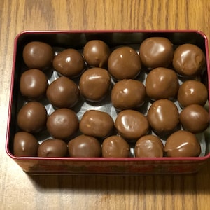 May include: A red tin box filled with chocolate candies. The candies are round and have a smooth, brown chocolate coating.