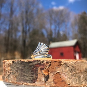 May include: A silver ring with a unique, fan-shaped design. The ring is sitting on a piece of wood with a blurred background of trees and a red building.