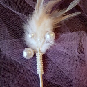 May include: A white feather with a string of white pearls attached to a white stick. The feather and pearls are on a purple tulle background.