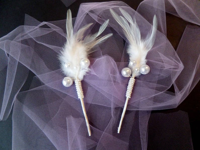 May include: Two white feather hair clips with pearl accents on a purple tulle background.