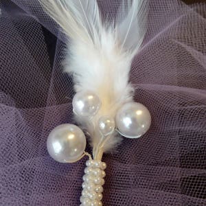 May include: A white feather with a pearl stem and white pearl accents. The feather and pearls are on a purple tulle background.