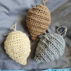 May include: Three crocheted ornaments in the shape of a pine cone. The ornaments are brown, beige, and gray. A blue measuring tape is in the foreground.