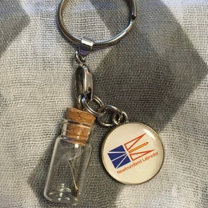 May include: A silver keychain with a small glass bottle containing a dandelion seed and a round metal charm with the flag of Newfoundland and Labrador and the text "Newfoundland Labrador".