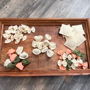 May include: A wooden tray with six different types of felt pasta shapes. The pasta shapes are white, green, and peach. The tray is on a wood floor.