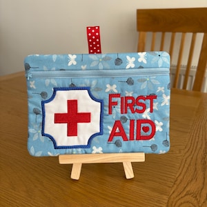 May include: A light blue fabric pouch with a zip closure and a red and white polka dot hanging loop. The pouch features a white cross with a red centre and the red embroidered text FIRST AID with a small heart replacing the letter I.