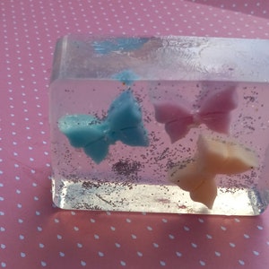 May include: A clear rectangular soap bar with three butterfly shapes embedded inside. The butterflies are blue, pink, and white. The soap bar is filled with silver glitter.