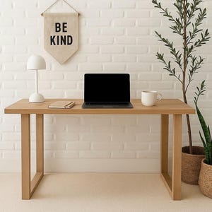 May include: A light wood desk with a laptop, notebook, and coffee mug. A banner above the desk reads "BE KIND". A white lamp and a potted plant are also on the desk. The desk is against a white brick wall.