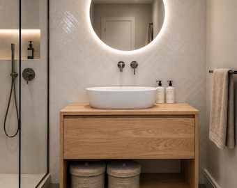May include: A bathroom vanity featuring a light oak wood cabinet with one drawer and an open bottom shelf containing two round woven baskets. A white oval vessel sink sits on top, paired with two dark metal wall-mounted faucets and a round LED mirror.