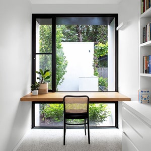 Könnte beinhalten: Ein heller Holzschreibtisch mit einem schwarzen Stuhl steht vor einem großen Fenster mit Blick auf einen üppigen grünen Garten. Der Schreibtisch ist an der Wand befestigt und hat eine weiße Oberfläche auf der rechten Seite.