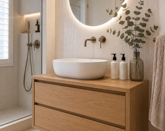 Puede incluir: Un mueble de baño flotante de roble claro con dos cajones, coronado por un lavabo ovalado blanco. Encima, un espejo redondo retroiluminado cuelga en una pared de azulejos blancos. Dos botellas con dosificador y un jarrón de cristal están encima.