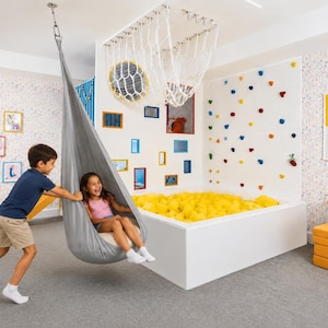 Op de afbeelding: Een grijze stoffen pods-schommel hangend aan het plafond, een witte ballenbak gevuld met gele ballen, een witte klimmuur met kleurrijke grepen, een wit plafondnet en een geel vloerkussen in een kamer met confetti-patroon behang.