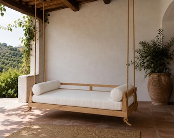 Può includere: Un divano letto sospeso rettangolare in legno, fissato con corde spesse a un soffitto in legno. Il divano è dotato di una struttura in legno naturale, un lungo cuscino rettangolare bianco e due cuscini cilindrici bianchi alle estremità.