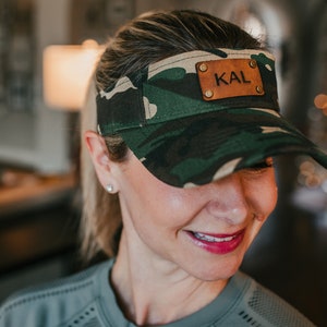 May include: A camouflage visor with a brown leather patch that says "KAL" on it.