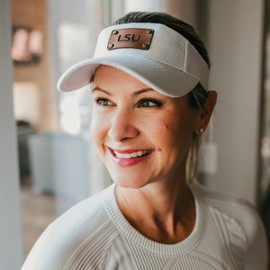 May include: A woman wearing a white visor with a brown leather patch that says "LSU" on it. She is smiling and looking to the right.
