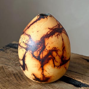Könnte beinhalten: Eine eiförmige Vase mit einer glatten, hellbraunen Oberfläche, die dunkle, verzweigte, blitzartige Muster aufweist. Die Vase hat eine kleine, kreisförmige Öffnung an der Oberseite, die einen dunklen Innenrand und eine abgerundete Form zeigt.