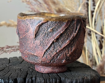 May include: A small, hand-thrown ceramic tea bowl with a rough, textured surface in shades of deep reddish-brown and charcoal black. The rim features a smooth, metallic gold-toned glaze, and the body has prominent, diagonal carved grooves.
