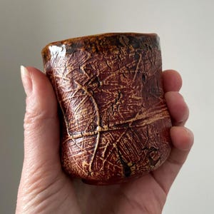 May include: A small, cylindrical ceramic cup with a deep reddish-brown glaze and a prominent, dark, crackled texture across the entire surface. The rim is slightly uneven, and the exterior features a network of raised, lighter-coloured lines.