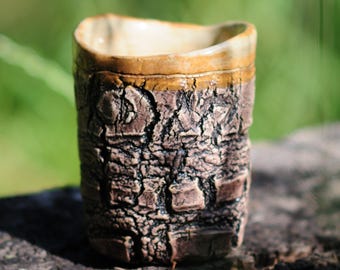 May include: A handcrafted ceramic cup with a textured, bark-like exterior in shades of brown and black. The rim is a lighter tan colour, and the interior is a creamy beige. The cup is designed for beverages.