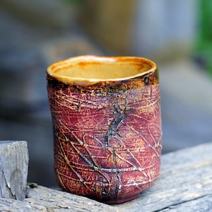 May include: A handmade ceramic tumbler with a textured, reddish-brown exterior and a golden-brown interior. The surface features intricate, etched patterns and a dark, crack-like design. The rim is a darker shade of brown.
