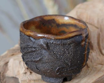 May include: A handmade, textured ceramic tea bowl with a dark brown exterior and a lighter brown rim. The bowl has a unique, organic shape and a small, rounded base. The interior is a warm, earthy brown color.
