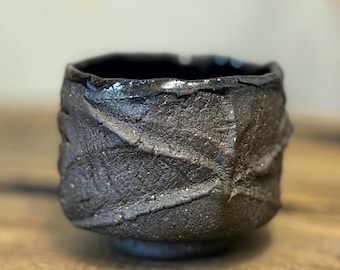 May include: A dark charcoal gray ceramic cup with a rough, textured exterior featuring raised leaf-like vein patterns. The rim is uneven and dark, with a deep, smooth interior. The vessel has a thick, sturdy base and a matte, earthy finish.
