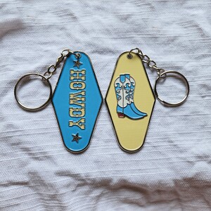 May include: Two metal keychains with a blue and yellow enamel finish. One keychain features the word "HOLLYWOOD" in gold lettering and the other keychain features a pair of blue cowboy boots with white accents.
