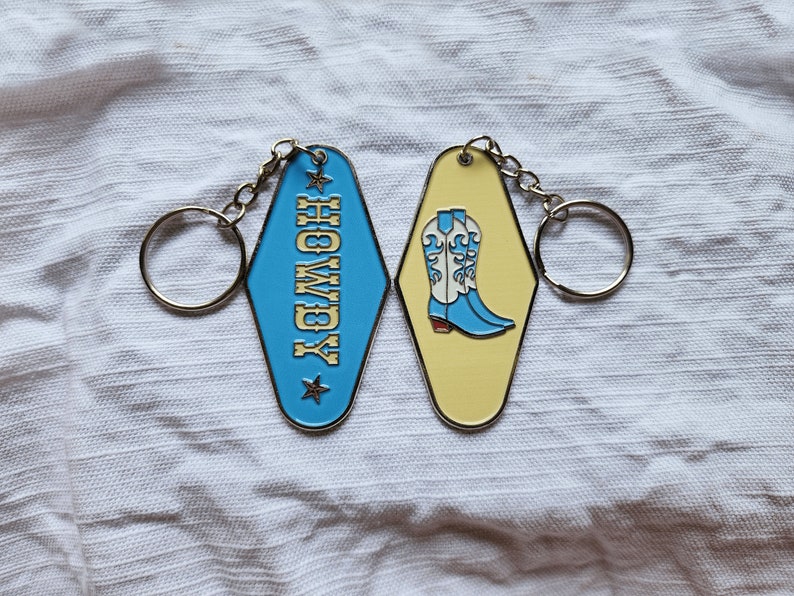 May include: Two metal keychains with a blue and yellow enamel finish. One keychain features the word "HOLLYWOOD" in gold lettering and the other keychain features a pair of blue cowboy boots with white accents.