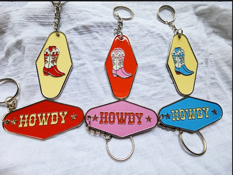 May include: Three keychains with a motel key design. Each keychain features a different colour: red, pink, and blue. The keychains have the word "HOWDY" written on them in gold lettering with a star on each side. Each keychain also features a pair of cowboy boots in a different colour: red, pink, and blue.