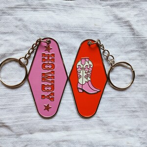 May include: Pink and orange motel keychains with a gold chain and ring. The pink keychain says "Howdy" with a star on each side. The orange keychain has a pair of pink cowboy boots with white tops.